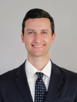 Smiling man with short black hair wearing a dark suit and tie.