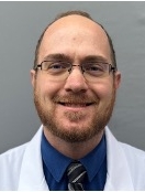 Smiling man with a light brown short beard and glasses wearing a white lab coat over a shirt and tie.