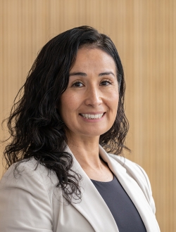 Smiling person with long wavy dark hair wearing a gray jacket over a charcoal top.