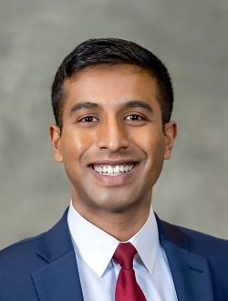 Smiling person with short black hair wearing a suit and tie.