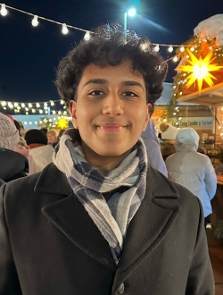 Smiling person with short black curly hair wearing a coat and scarf.