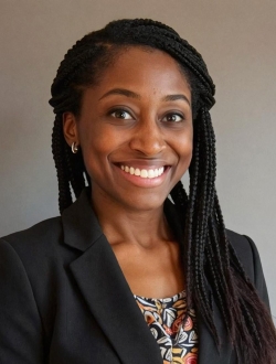 Perspn with long braids, smiling and wearing a black jacket over a multicolored top.