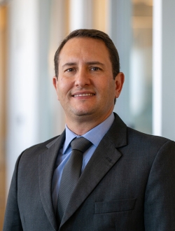Person with short dark brown hair wearing a dark suit, blue shirt and tie.