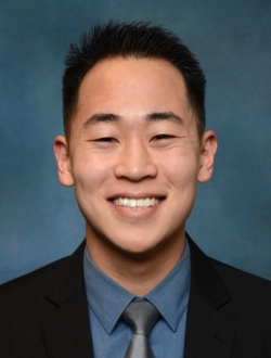 Smiling person with short black hair wearing a suit and tie.