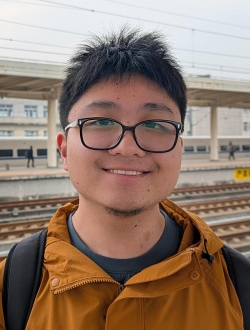 Smiling person with short spiky black hair wearing glasses and an orange jacket.