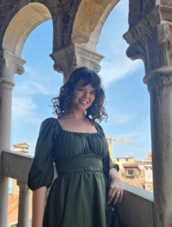Person with long curly brown hair wearing a green dress and standing on an open stone staircase.