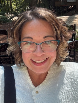 Person with shoulder length light brown hair, smiling, wearing glasses, and a white jacket.