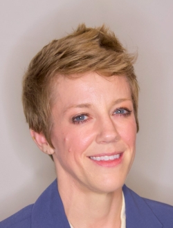 Smiling person with short blonde hair wearing a blue jacket.