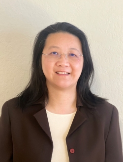 Smiling person with shoulder-length black hair wearing a brown jacket over a light colored top and glasses.