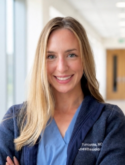 Smiling person with long blonde hair wearing a navy blue jacket over scrubs.