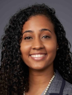 Smiling person with long black curly hair wearing a suit jacket and pearls.