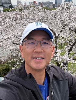 Smiling person wearing glasses a white baseball cap and a black windbreaker standing in front of a group of cherry trees in full blossom.