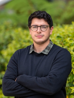 Smiling person with short dark hair wearing a dark sweater over a shirt with greenery in the background.