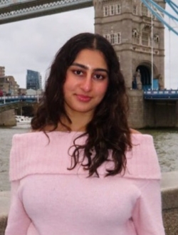 person with long brown hair wearing a pink sweater standing in front of London Bridge.