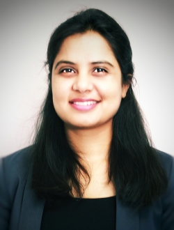 Smiling person with long black hair wearing a dark green jacket over a black top.