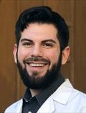 Man smiling with dark hair and beard wearing a white lab coat over a collared shirt.