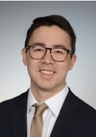 Smiling man with short black hair and glasses wearing a suit and tie.