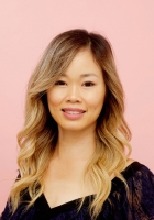 Woman with long light brown hair wearing a dark top in front of a pink background.