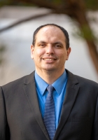 Smiling man with short black hair wearing a dark suit with a blue tie and shirt with a tree and building in the background.