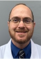 Smiling man with a light brown short beard and glasses wearing a white lab coat over a shirt and tie.