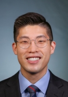 Smiling person with short black hair wearing glasses and a suit and tie.