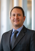 Person with short dark brown hair wearing a dark suit, blue shirt and tie.