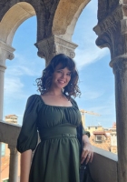 Person with long curly brown hair wearing a green dress and standing on an open stone staircase.