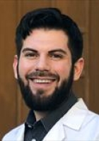 Man smiling with dark hair and beard wearing a white lab coat over a collared shirt.